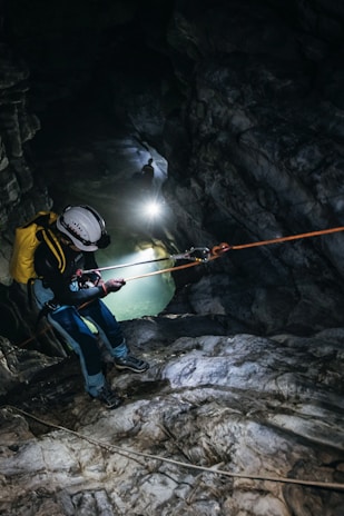 A person is engaged in a caving or rappelling activity inside a rocky cave. They are equipped with climbing gear including a helmet and ropes. The cave is dark, illuminated by the light from the person, highlighting the rugged textures of the stones and the presence of a body of water in the background.