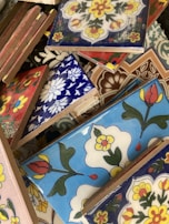 A variety of colorful ceramic tiles with intricate floral and geometric patterns. The tiles are stacked haphazardly, showcasing designs in blue, red, yellow, and white with bold outlines. Each tile features distinct artistic motifs that reflect a blend of traditional and modern styles.