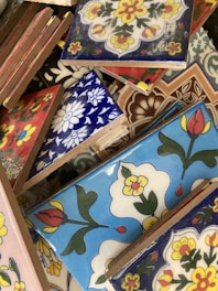 A variety of colorful ceramic tiles with intricate floral and geometric patterns. The tiles are stacked haphazardly, showcasing designs in blue, red, yellow, and white with bold outlines. Each tile features distinct artistic motifs that reflect a blend of traditional and modern styles.