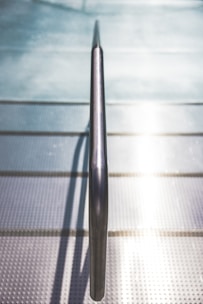 A metallic handrail is centrally aligned, stretching horizontally across a textured surface. The background reveals a grid-like pattern with reflections of sunlight creating a bright, luminous effect. The setting suggests a modern architectural design with an emphasis on industrial aesthetics.