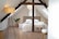 A cozy attic bedroom features a bed with white linens and a neutral headboard. The space is accented by exposed dark wooden beams. Warm lighting comes from a modern floor lamp with a cylindrical shade and a small bedside lamp. There's a potted plant adding a touch of greenery beside a small table. The room has a wooden floor and two small wooden stools at the foot of the bed.