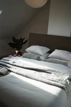 A neatly made bed with a thick, light-colored blanket and two white pillows is positioned against a neutral-toned wall. A soft, warm light from a lamp on a bedside table creates a cozy atmosphere. A large green plant adds freshness to the room, and an open book lies on the blanket near the pillows.