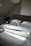 A neatly made bed with a thick, light-colored blanket and two white pillows is positioned against a neutral-toned wall. A soft, warm light from a lamp on a bedside table creates a cozy atmosphere. A large green plant adds freshness to the room, and an open book lies on the blanket near the pillows.