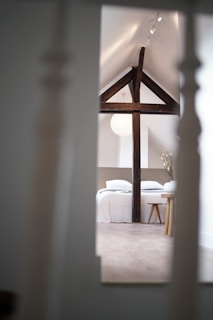 A cozy bedroom located in an attic space featuring exposed dark wooden beams across a peaked ceiling. The room contains a neatly made bed with white linens, and a small wooden stool beside it. Soft lighting enhances the warm ambiance, and a pendant lamp hangs from the ceiling. A minimalistic interior design approach is evident, with a focus on natural materials and neutral tones.