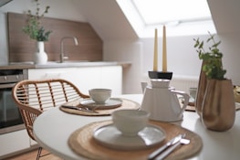 A cozy kitchen setup featuring a round table with place settings, including plates, bowls, and cutlery on woven placemats. There are two lit candles in a holder, a white teapot, and vases with green plants. The background shows a modern kitchen with a sink and a wood-paneled backsplash under a skylight.