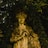 An aged stone statue of a woman with curly hair stands amidst dense, dark green foliage. The figure is detailed, holding a globe-like object and features a serene expression. The statue appears weathered and coated with patches of moss and lichen, blending organically with its natural surroundings.