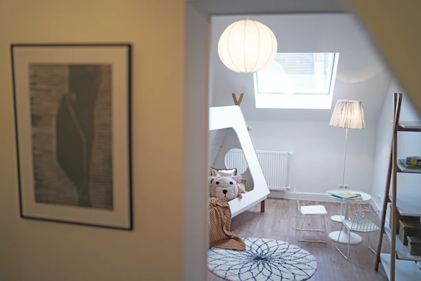 A cozy, modern children's room with a playful tent-like bed and a bear-themed pillow. The room is illuminated by natural light from a skylight and a standing lamp with a paper shade. A small chair and table set is placed nearby, with a simple ladder bookshelf against the wall. The decor features neutral tones with wooden flooring and a round rug with a circular pattern.