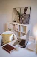 A modern office space featuring a white table with a laptop, a closed notebook, and a coffee mug. A white chair with a shiny gold pillow is in front of the desk. In the background, there is a white bookshelf with neatly arranged folders and two decorative framed photo prints resting on top. A pair of grey vases with a single green leaf add a touch of greenery.