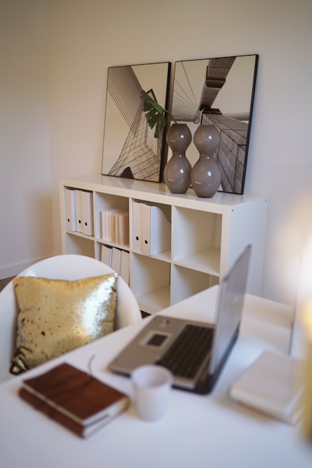 A modern office space featuring a white table with a laptop, a closed notebook, and a coffee mug. A white chair with a shiny gold pillow is in front of the desk. In the background, there is a white bookshelf with neatly arranged folders and two decorative framed photo prints resting on top. A pair of grey vases with a single green leaf add a touch of greenery.