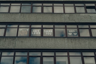 A series of windows on a concrete building displaying messages of gratitude and support, including phrases like 'Stay Safe,' 'Save Lives,' 'Thank You,' and 'NHS' accompanied by hearts.