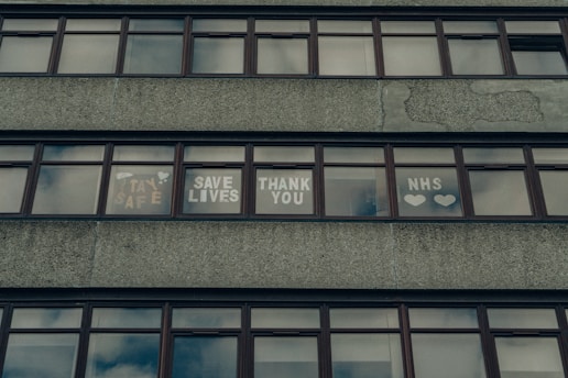 A series of windows on a concrete building displaying messages of gratitude and support, including phrases like 'Stay Safe,' 'Save Lives,' 'Thank You,' and 'NHS' accompanied by hearts.
