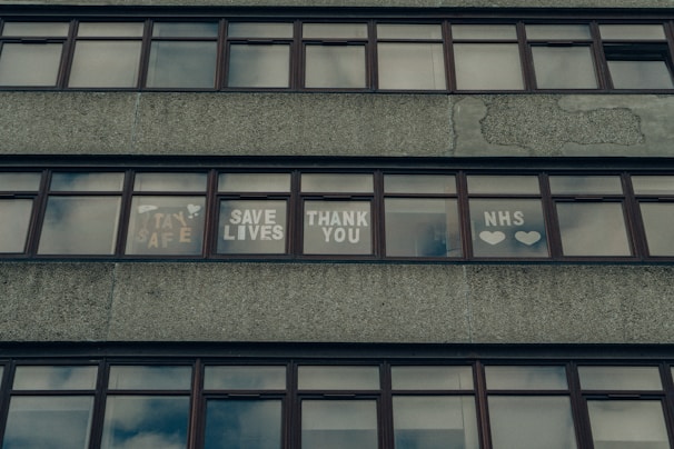 A series of windows on a concrete building displaying messages of gratitude and support, including phrases like 'Stay Safe,' 'Save Lives,' 'Thank You,' and 'NHS' accompanied by hearts.