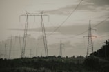 A landscape featuring several tall electrical transmission towers standing amidst an open field. The towers are interconnected by numerous power lines. The sky is overcast with a mixture of grey clouds, adding to the subdued lighting of the scene. Vegetation and distant trees line the horizon, providing a contrasting natural element to the industrial structures.