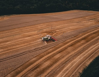 Aerial view of a large wheat field with a combine harvester moving across the land, leaving a trail of harvested crop. The field has long, parallel rows, and there is a dense forest at the edge of the field.