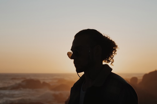 A silhouette of a person with curly hair and glasses standing in front of a calm ocean at sunset. The sky is lit with warm hues of orange and yellow, creating a serene backdrop.