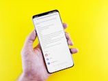 A hand holding a smartphone with a screen displaying a settings menu in Russian, likely related to account or device settings. The phone is against a bright yellow background.