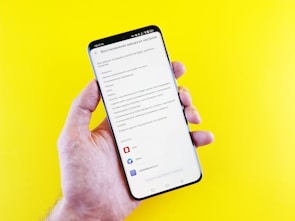 A hand holding a smartphone with a screen displaying a settings menu in Russian, likely related to account or device settings. The phone is against a bright yellow background.