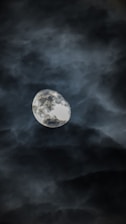 A luminous moon is partially shrouded by dark, swirling clouds. The clouds form a dramatic backdrop, creating a mysterious and atmospheric scene.