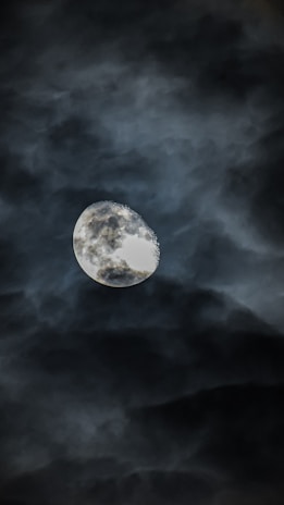 A luminous moon is partially shrouded by dark, swirling clouds. The clouds form a dramatic backdrop, creating a mysterious and atmospheric scene.