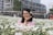 A woman stands in a vibrant field of white daisies, smiling and posing with a flower tucked behind her ear. She is wearing glasses and a pink shirt. In the background, other people are visible, and there is a partially constructed building surrounded by greenery.