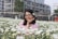 A woman stands in a vibrant field of white daisies, smiling and posing with a flower tucked behind her ear. She is wearing glasses and a pink shirt. In the background, other people are visible, and there is a partially constructed building surrounded by greenery.
