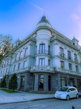 A three-story historic building with white and beige tones, featuring arched windows, and a rounded corner section with the word 'BATUM' inscribed on it. Balconies adorn each floor, adding to the architectural elegance. The setting is urban with cobblestone streets and a few parked cars. A tree and some bushes are visible near the sidewalk, providing a touch of greenery.