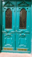 A pair of ornate teal double doors featuring intricate wooden carvings and decorative molding. The doors have metal grid windows and brass handles, adding to the classic design.