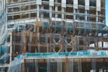 A partially constructed building covered in mesh netting likely for safety and dust control. The structure is primarily made of steel beams and partially visible cement floors. Scaffolding is present along the edges and construction materials are stacked at different levels.