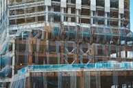 A partially constructed building covered in mesh netting likely for safety and dust control. The structure is primarily made of steel beams and partially visible cement floors. Scaffolding is present along the edges and construction materials are stacked at different levels.