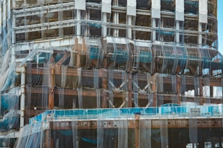 A partially constructed building covered in mesh netting likely for safety and dust control. The structure is primarily made of steel beams and partially visible cement floors. Scaffolding is present along the edges and construction materials are stacked at different levels.