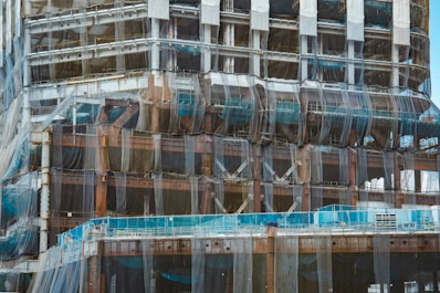 A partially constructed building covered in mesh netting likely for safety and dust control. The structure is primarily made of steel beams and partially visible cement floors. Scaffolding is present along the edges and construction materials are stacked at different levels.