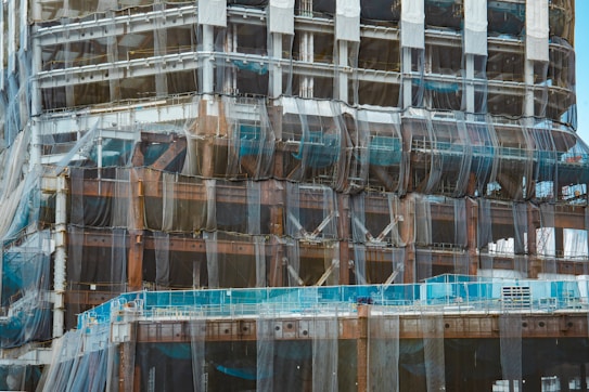 A partially constructed building covered in mesh netting likely for safety and dust control. The structure is primarily made of steel beams and partially visible cement floors. Scaffolding is present along the edges and construction materials are stacked at different levels.