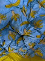 A crumpled bed sheet featuring a playful pattern of cartoon bananas on a bright blue background. The bananas are stylized, some of them whole and some peeled, with a simple design that emphasizes their yellow hue. The sheet contrasts sharply with a small visible part of a solid yellow fabric beneath.