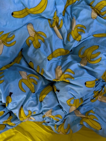 A crumpled bed sheet featuring a playful pattern of cartoon bananas on a bright blue background. The bananas are stylized, some of them whole and some peeled, with a simple design that emphasizes their yellow hue. The sheet contrasts sharply with a small visible part of a solid yellow fabric beneath.