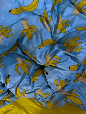 A crumpled bed sheet featuring a playful pattern of cartoon bananas on a bright blue background. The bananas are stylized, some of them whole and some peeled, with a simple design that emphasizes their yellow hue. The sheet contrasts sharply with a small visible part of a solid yellow fabric beneath.
