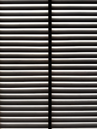 Horizontal blinds partially open with light filtering through the slats, casting striped shadows and creating a geometric pattern.