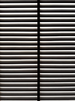 Horizontal blinds partially open with light filtering through the slats, casting striped shadows and creating a geometric pattern.