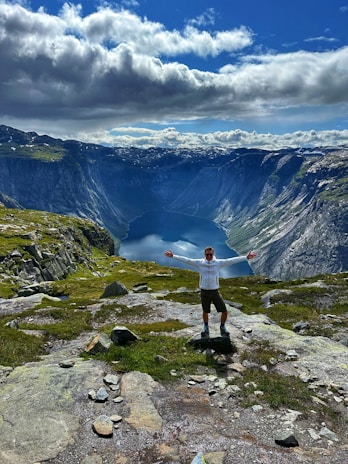 A person, dressed in a white jacket and shorts, stands with arms outstretched on rocky terrain overlooking a deep blue fjord surrounded by high, steep cliffs. In the background, a partly cloudy sky contrasts with the green and rocky mountain landscape.