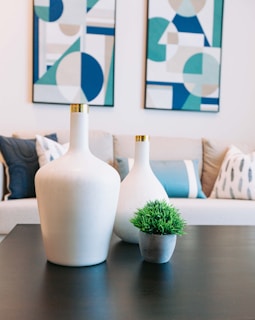Two white vases with gold accents and a small green potted plant are on a dark coffee table. Behind them, there is a beige couch with blue and white pillows. Two modern, geometric art pieces in shades of blue, teal, and beige are hung on the wall.