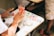 A person is seated at a desk, engaged in painting activities. The desk is scattered with various small containers of colorful paint. The person has their hands poised above the desk, possibly waiting for paint to dry or mixing colors. A plastic cup with a straw and orange beverage is visible nearby.