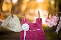 A collection of crocheted baby clothing items are hanging on a clothesline. The items include a colorful crochet hat with a flower adornment, a pink crochet dress featuring a white flower, and a pair of pink and white crochet baby sandals with flower decorations. The background is softly blurred and bathed in warm, golden light, creating a serene outdoorsy setting.