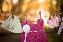A collection of crocheted baby clothing items are hanging on a clothesline. The items include a colorful crochet hat with a flower adornment, a pink crochet dress featuring a white flower, and a pair of pink and white crochet baby sandals with flower decorations. The background is softly blurred and bathed in warm, golden light, creating a serene outdoorsy setting.