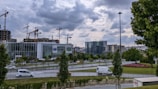 A cityscape with several construction cranes towering above buildings in progress against a cloudy sky. A modern glass building sits prominently in the foreground, surrounded by greenery and a few vehicles on the roads. The scene suggests ongoing urban development.