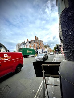 A bustling street scene with vehicles, including a red Royal Mail van, a green Harrods truck, and several cars. The background features a prominent brick building with ornate architectural details and multiple chimneys. The sky is overcast, adding a subdued ambiance to the setting.