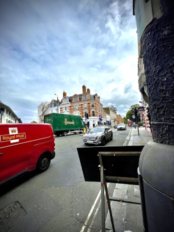 A bustling street scene with vehicles, including a red Royal Mail van, a green Harrods truck, and several cars. The background features a prominent brick building with ornate architectural details and multiple chimneys. The sky is overcast, adding a subdued ambiance to the setting.