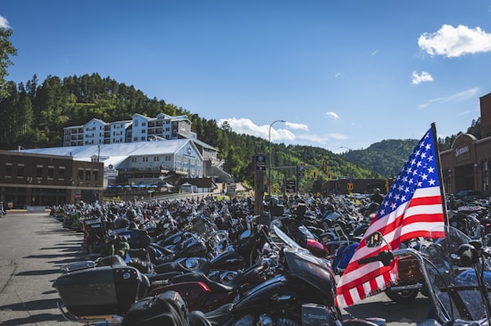 A parking lot is filled with numerous motorcycles, creating a dense sea of bikes. An American flag is prominently displayed among the motorcycles. In the background, there are buildings with a wooded hillside rising behind them under a clear blue sky.