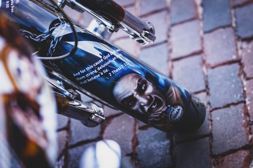 A close-up of a motorcycle part features a detailed illustration of a sinister face, possibly depicting a mythical or fictional character. Above the image, a biblical verse from 2 Thessalonians 2:11 is visible. The background consists of a paved stone surface.