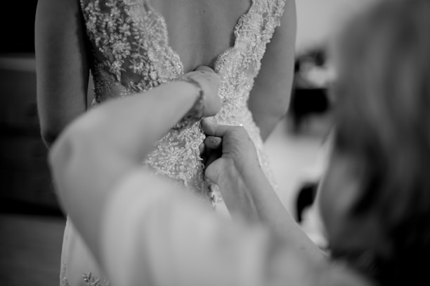 A close-up of a person fastening the intricate buttons on the back of an embroidered wedding dress. The gown features delicate lace patterns and embellishments. The scene is captured in black and white, emphasizing the texture and details.