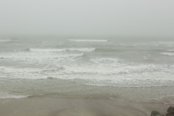 Gray, overcast skies hang above turbulent ocean waves crashing towards a sandy shore. The atmosphere feels misty and foggy, with reduced visibility at the horizon.