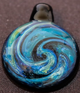 A handcrafted glass pendant with intricate swirling patterns resembling a blue and green galaxy or whirlpool, set against a dark background. The design includes shimmering elements that add a subtle sparkle.